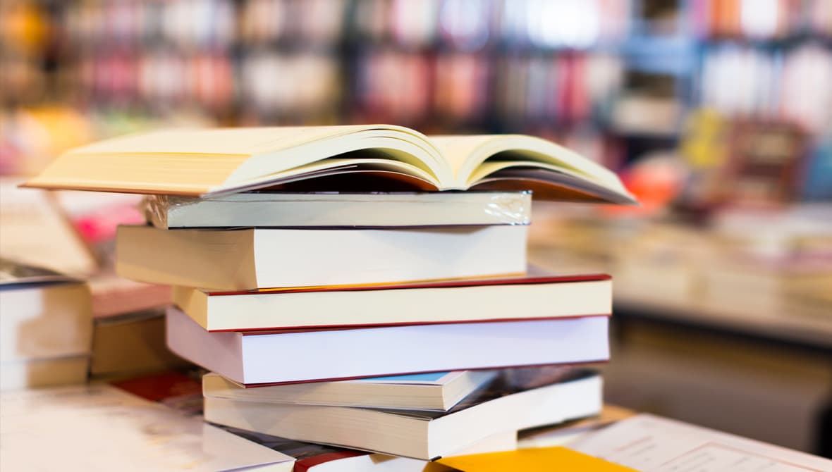 A pile of books on a table, with bookshelves in the background.